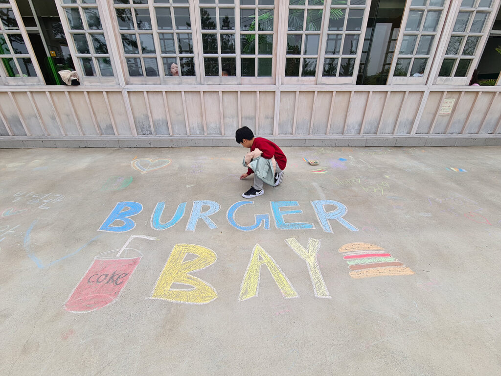 Child drawing colorful chalk art of “Burger Bay” with a burger and soda on the ground.