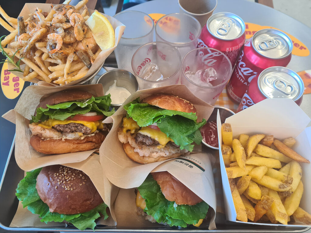 Tray of Golden Bay Burgers served with fries, shrimp-topped fries, and Coca-Cola cans.