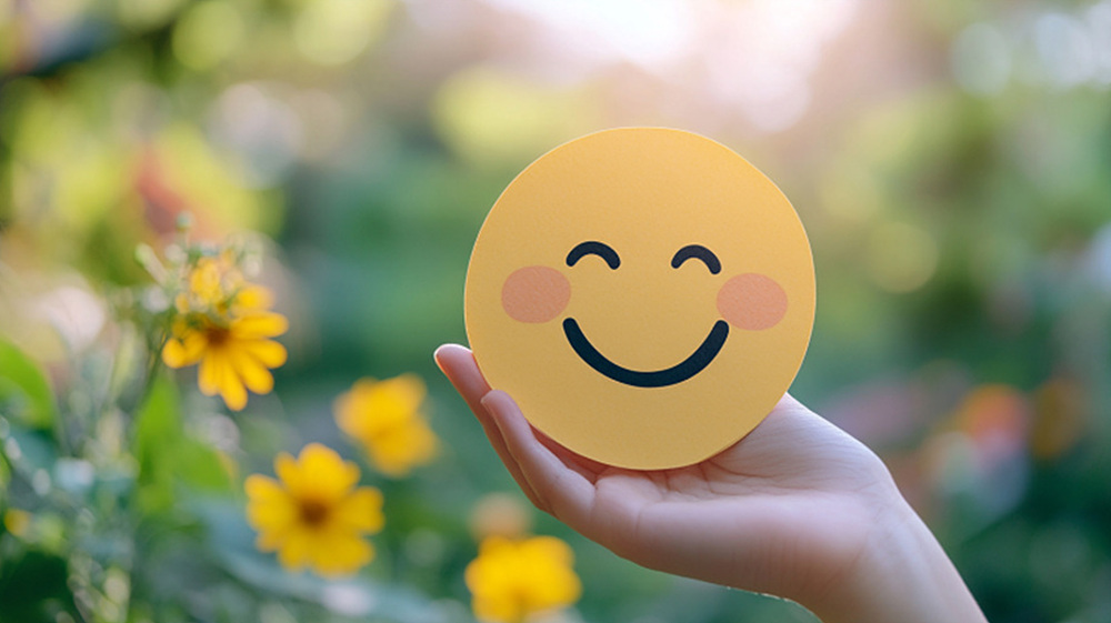 A hand holding a yellow smiley face in a flower field.